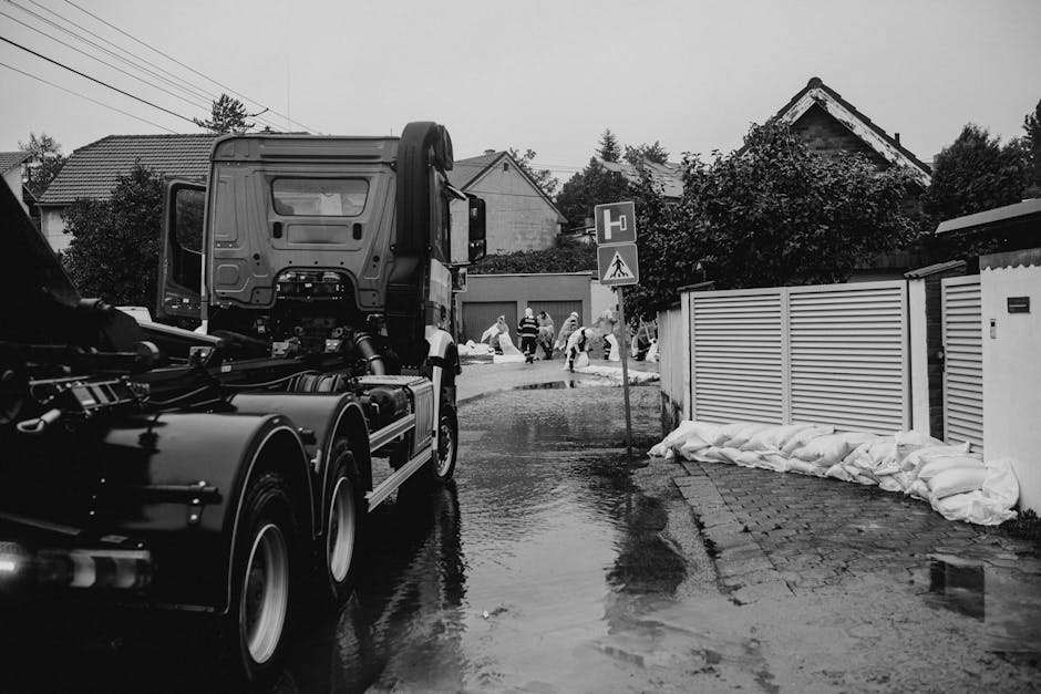 Image of a residential street with several houses and trees, where a team of workers dressed in yellow and orange protective suits are engaged in emergency cleaning and stain removal. Some individuals